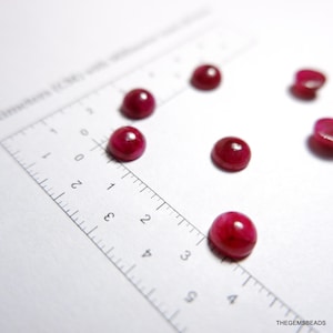 May include: Several polished, round, deep red gemstones are scattered on a white surface next to a ruler. The gemstones are cabochons, and the ruler is marked in centimetres.