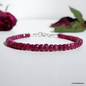 May include: A ruby gemstone bracelet. The bracelet features small, faceted, deep red ruby beads strung together with a silver clasp. The beads are a rich, saturated color. The background is white with a blurred rose.