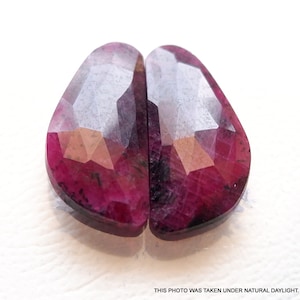 May include: Two faceted, pink ruby gemstones with a dark red hue and a clear, glassy finish. The stones are cut in a teardrop shape and are set side by side.
