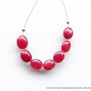 May include: A strand of oval-shaped, red gemstone beads strung on a silver wire. The beads are smooth and polished.