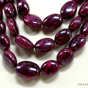 Pode incluir: Um fio de contas de rubi vermelho escuro, de forma oval e polidas, com um acabamento liso e brilhante. As contas estão dispostas em três filas, criando um efeito em camadas.