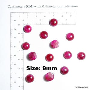 Op de afbeelding: Een verzameling ronde, gepolijste, magenta-kleurige kralen, elk ongeveer 9 mm in diameter, ligt verspreid op een witte ondergrond. Een liniaal met centimeter- en millimeter-markeringen is zichtbaar. De tekst "Size: 9mm" is aanwezig.