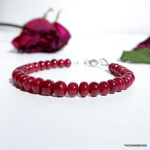 Puede incluir: Una pulsera de cuentas rojas con un cierre plateado. Las cuentas son de un color rojo intenso y rico y parecen pulidas. La pulsera se muestra sobre una superficie blanca, con un fondo borroso con una rosa roja oscura.