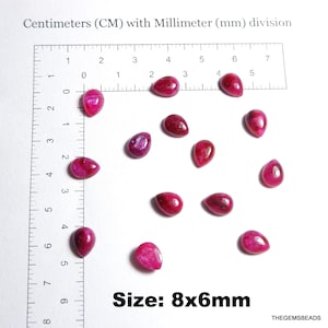 Op de afbeelding: Een verzameling druppelvormige, gepolijste, dieproze edelstenen, elk ongeveer 8x6mm groot, verspreid op een wit oppervlak. Een liniaal met centimeter- en millimeter-markeringen is op de achtergrond. De tekst "Size: 8x6mm" is zichtbaar.