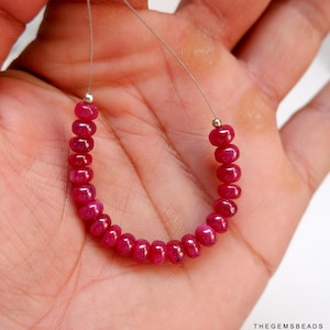 May include: A strand of deep red ruby beads strung on a silver wire. The beads are round and smooth, and they are arranged in a u-shape.
