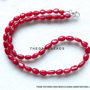 May include: A strand of red gemstone beads with a silver clasp. The beads are oval shaped and have a smooth, polished finish. TheGEMSBeads is printed on the white background.
