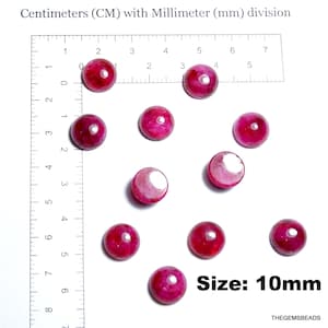 May include: A collection of ten round, polished, deep pink gemstones, each approximately 10mm in diameter, displayed on a white surface next to a ruler marked in centimeters. The text "Size: 10mm" is visible.