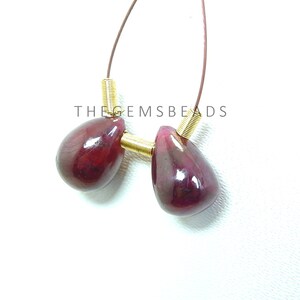 May include: Two teardrop-shaped ruby gemstones with a gold-toned connector. The gemstones are a deep red color and have a glossy finish. The connector is a small, round piece of metal with a smooth surface.