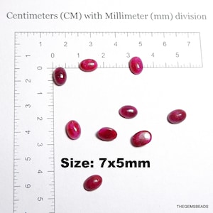 Peut inclure: Plusieurs pierres précieuses ovales, rouge foncé, d'environ 7 mm sur 5 mm, sont disposées sur une surface blanche, à côté d'une règle graduée en centimètres et millimètres. Le texte "Size: 7x5mm" est visible.