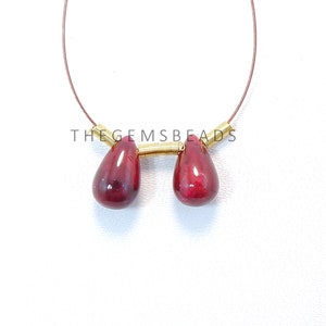 May include: Two teardrop-shaped red gemstone beads strung on a gold wire with a gold wire loop at the top. The beads are connected by a gold wire coil. The text "THE GEMSBeads" is visible below the beads.