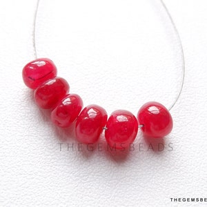 May include: A string of six bright red, polished gemstone beads. The oval-shaped beads are strung on a thin, clear wire. The gemstones have a glossy finish and are set against a white background. The text "THEGEMSBEADS" is visible.