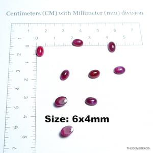 May include: Oval-shaped, deep red gemstones are scattered across a white surface, with a ruler in the background. The text "Size: 6x4mm" is displayed. The gemstones are approximately 0.24 x 0.16 inches in size.