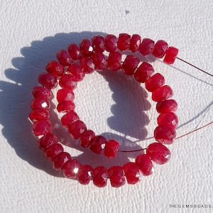 May include: A strand of deep red faceted gemstone beads. The beads are strung on a thin wire and are arranged in a circular pattern. The beads are all approximately the same size and shape.