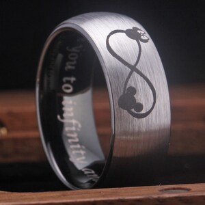 May include: A silver tungsten carbide ring with a brushed finish. The ring features an infinity symbol with two Mickey Mouse heads inside. The inscription "You to infinity and" is engraved on the inside of the ring.