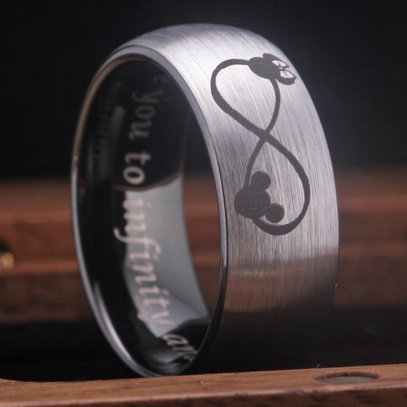 May include: A silver tungsten carbide ring with a brushed finish. The ring features an infinity symbol with two Mickey Mouse heads inside. The inscription "You to infinity and" is engraved on the inside of the ring.
