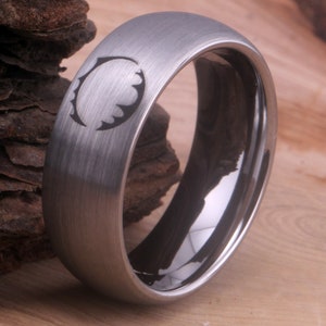 Op de afbeelding: Een geborstelde zilveren ring met een zwarte binnenband en een gestileerd ontwerp aan de bovenkant. Het ontwerp is een cirkel met een gekartelde rand.