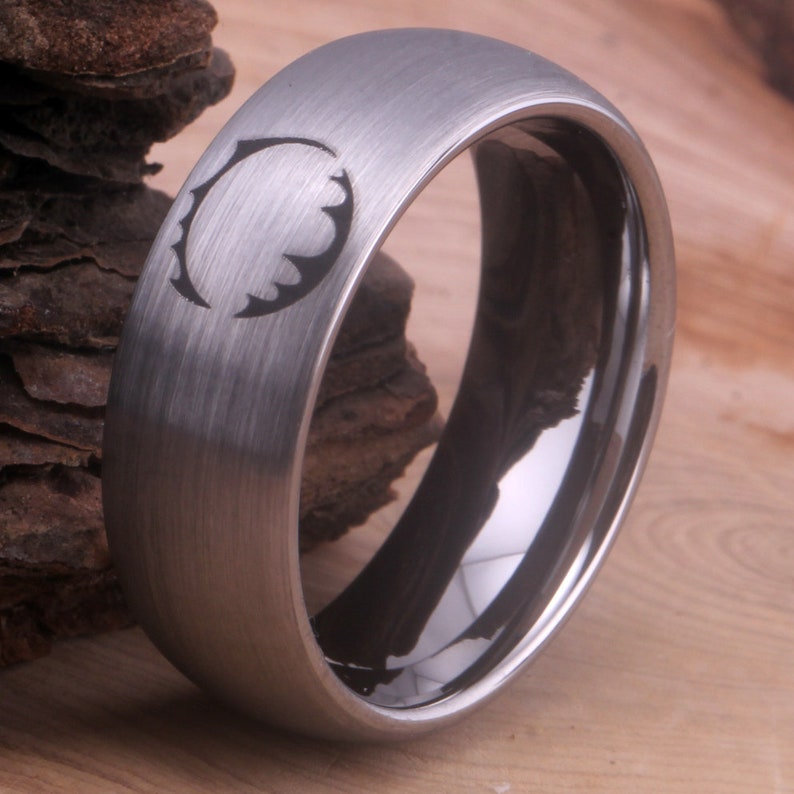 Op de afbeelding: Een geborstelde zilveren ring met een zwarte binnenband en een gestileerd ontwerp aan de bovenkant. Het ontwerp is een cirkel met een gekartelde rand.