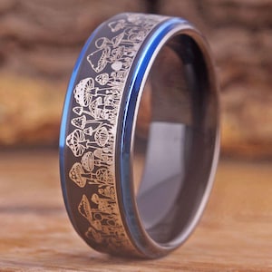 Puede incluir: Un anillo de carburo de tungsteno negro con un borde azul y un diseño de hongo plateado grabado alrededor de la banda.
