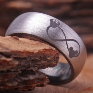 May include: A silver tungsten carbide ring with a brushed finish. The ring features an infinity symbol with a Mickey Mouse head at the top and three Mickey Mouse heads at the bottom.