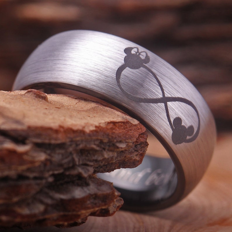 May include: A silver tungsten carbide ring with a brushed finish. The ring features an infinity symbol with a Mickey Mouse head at the top and three Mickey Mouse heads at the bottom.
