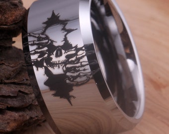 May include: A silver tungsten carbide ring with a black skull design engraved on the band. The skull is surrounded by a design of hands reaching out from behind it.