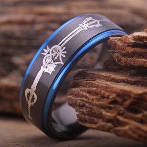 May include: A black and blue tungsten carbide ring with a silver engraved design of a keyblade and heart. The ring is on a wooden surface.