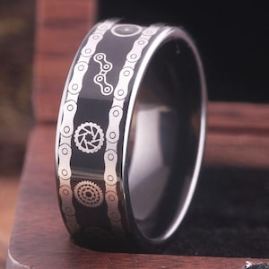 Puede incluir: Un anillo de carburo de tungsteno negro con un diseño de cadena y engranajes plateados.