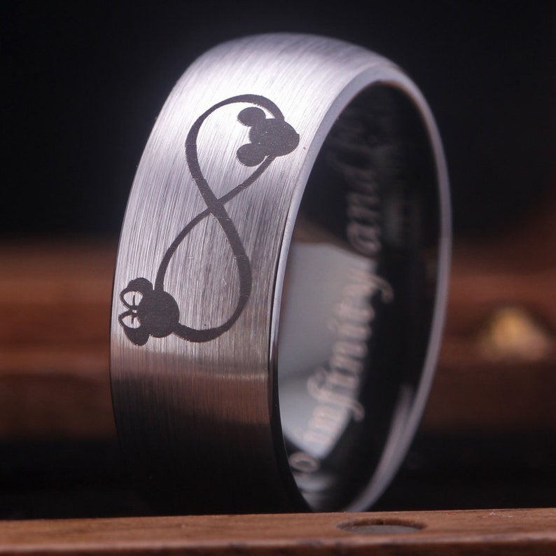 May include: A silver tungsten carbide ring with an infinity symbol and two Mickey Mouse heads engraved on the band. The ring is brushed and has a black inner band.