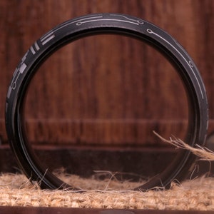 May include: A black ring with a silver circuit board design. The ring is sitting on a brown surface.