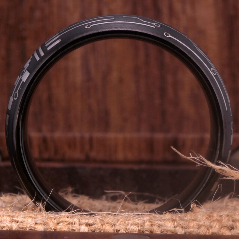 May include: A black ring with a silver circuit board design. The ring is sitting on a brown surface.