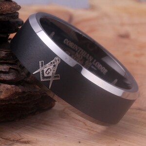 May include: A black tungsten carbide ring with a silver beveled edge. The ring has a Masonic symbol engraved on the top and the words "Corinthian Lodge No. 69" engraved on the side.