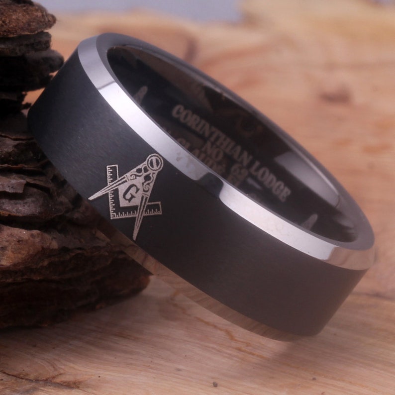 May include: A black tungsten carbide ring with a silver beveled edge. The ring has a Masonic symbol engraved on the top and the words "Corinthian Lodge No. 69" engraved on the side.