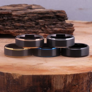 May include: Four black tungsten carbide rings with different colored trim. The rings are stacked on top of each other on a wooden surface. The rings have a rose gold, blue, and silver trim.