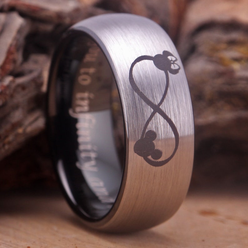 May include: A silver tungsten carbide ring with a brushed finish. The ring features an infinity symbol with two Mickey Mouse heads inside. The inside of the ring is engraved with the words "Forever Family".