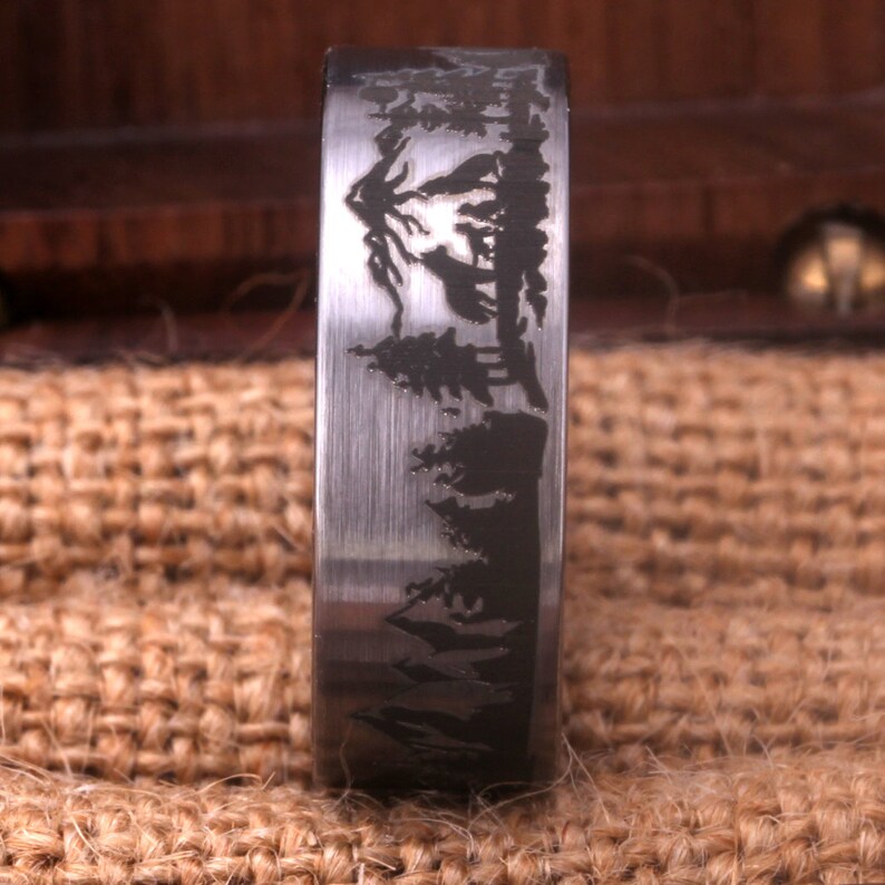 May include: A silver ring with a brushed finish and an engraved design of a mountain range and trees.