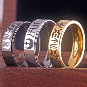 Könnte beinhalten: Drei Ringe in verschiedenen Farben mit eingravierten Star Wars-Symbolen und Text. Die Ringe sind schwarz, silber und gold.
