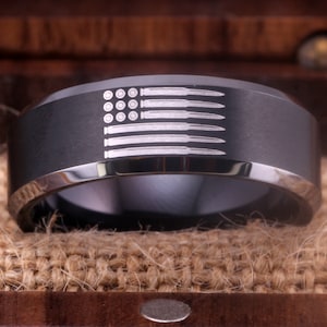 Puede incluir: Un anillo de carburo de tungsteno negro con un diseño grabado con láser de una bandera estadounidense hecha de balas.