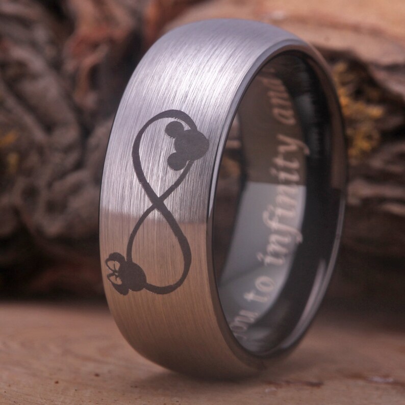 May include: A silver tungsten carbide ring with a brushed finish. The ring features an infinity symbol with two Mickey Mouse heads inside. The inside of the ring is engraved with the text "From to infinity and beyond".