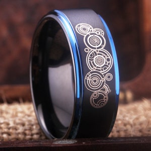 May include: A black and blue tungsten carbide ring with a silver design of a series of concentric circles.