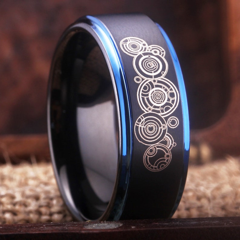 May include: A black and blue tungsten carbide ring with a silver design of a series of concentric circles.