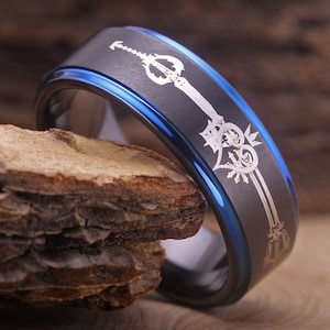 May include: A black and blue tungsten carbide ring with a silver engraved design of a keyblade and heart.