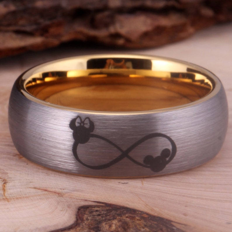 May include: A gray and gold wedding band with an infinity symbol and two Mickey Mouse heads engraved on the band.