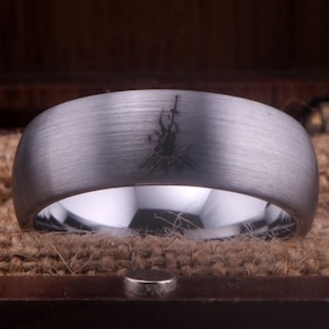 Op de afbeelding: Een zilveren ring van wolfraamcarbide met een geborstelde afwerking en een zwart gegraveerd ontwerp van een kampvuur met een zwaard.