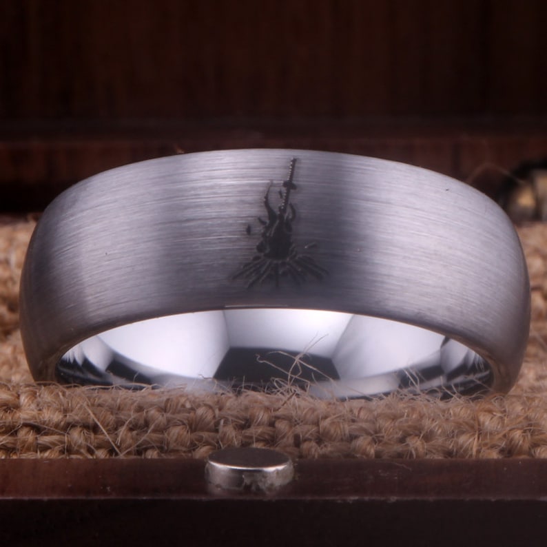 Op de afbeelding: Een zilveren ring van wolfraamcarbide met een geborstelde afwerking en een zwart gegraveerd ontwerp van een kampvuur met een zwaard.