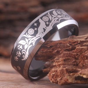 May include: A black tungsten carbide ring with a silver wolf design engraved on the band. The wolf is surrounded by a floral pattern and stars. The ring is on a wooden surface.