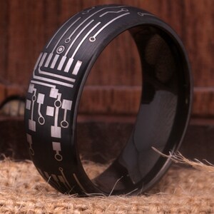 May include: A black tungsten carbide ring with a silver circuit board design. The ring is polished and has a brushed finish.