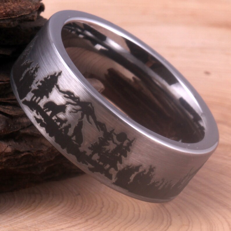 May include: A silver ring with a brushed finish and a black silhouette of a mountain range and trees engraved on the band.