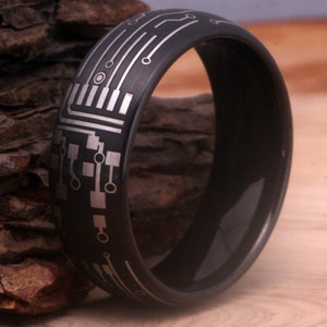 May include: A black tungsten carbide ring with a silver circuit board design engraved on the band.