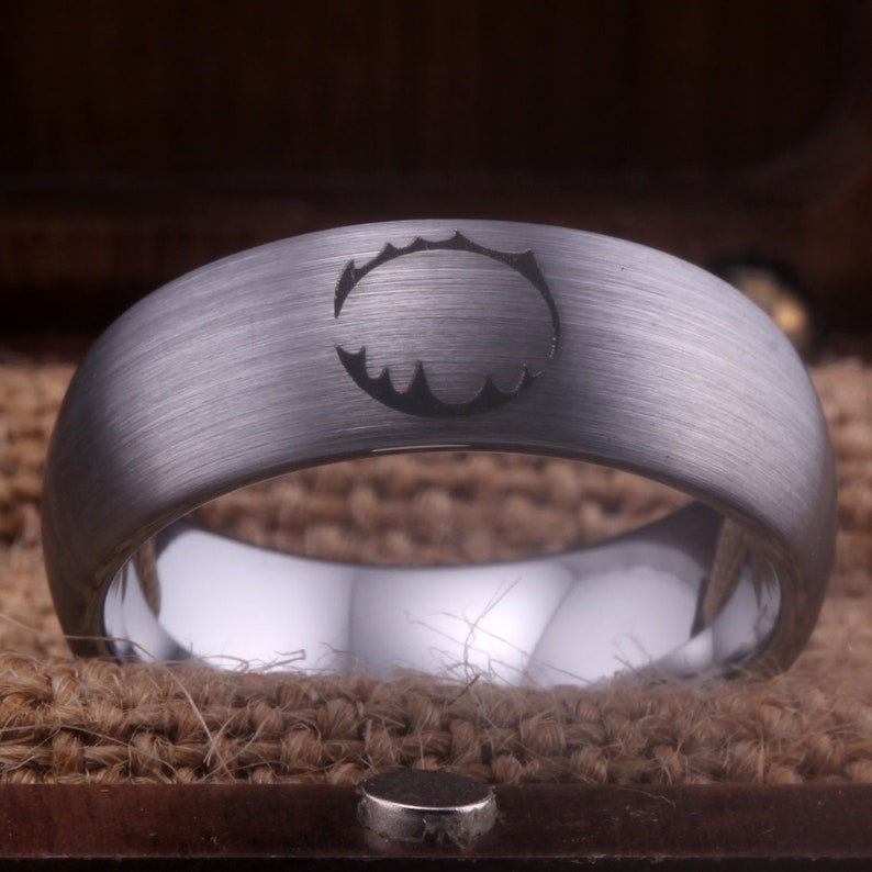 Op de afbeelding: Een geborstelde zilveren ring van wolframcarbide met een zwart gegraveerd ontwerp. Het ontwerp is een cirkel met een gekartelde rand.