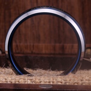 May include: A blue and silver tungsten carbide ring with a decorative design. The ring is shown from the side, with the inside of the ring facing the camera.
